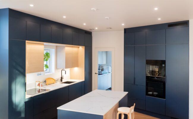 Avokeittion-spottivalaistus-on-aseteltu-kehaan-kaapistojen-eteen_LedStorefi_ - LedStore Tyylikäs keittiö, jossa on tummansiniset kaapistot ja valkoinen marmoritaso. Tilassa on ikkuna, jonka vieressä on viherkasvi ruukussa, sekä induktioliesi, uuni ja pieni puinen jakkara. Upotettu LED-valaistus valaisee tilan kirkkaasti. Tämä kaikki löytyy LedStore.fi:n laajasta LED-valaistusvalikoimasta!