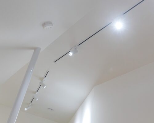 Korkean-tilan-ratkaisuna-toimivat-FOKUS-kiskovalot_LedStorefi_ - LedStore Katto, jossa on valkoinen, minimalistinen muotoilu ja useita LED-kiskovalaisimia, tulee yksi loistaa kirkkaasti. Kattoon on kiinnitetty myös pieni, pyöreä palovaroitin. Huone vaikuttaa moderniilta ja siistiltä. Tämä sopii hyvästi. LedStorefi:n laaduseen led-valaistusvalikoimaan.