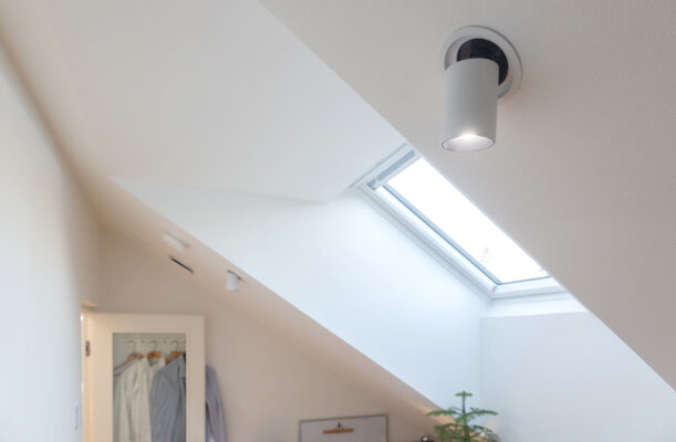 Tyohuoneen-jyrkan-katon-valaisussa-KOLO-spotit-ovat-oikeassa-paikassa_LedStorefi_ - LedStore Minimalistinen huone vinolla katolla on valaistu pyöreällä LED-kattovalaisimella sekä kattoikkunalla, valikoimaa löytyy LedStore.fi-verkkokaupasta. Kattoikkunan alla sijaitsee pieni kasvi. Vasemmalla seinällä on osittain näkyvissä ovi, josta roikkuu vaatteita. Valkoiset seinät korostavat huomioita sisustusta ja luovatin ilmapiirin.