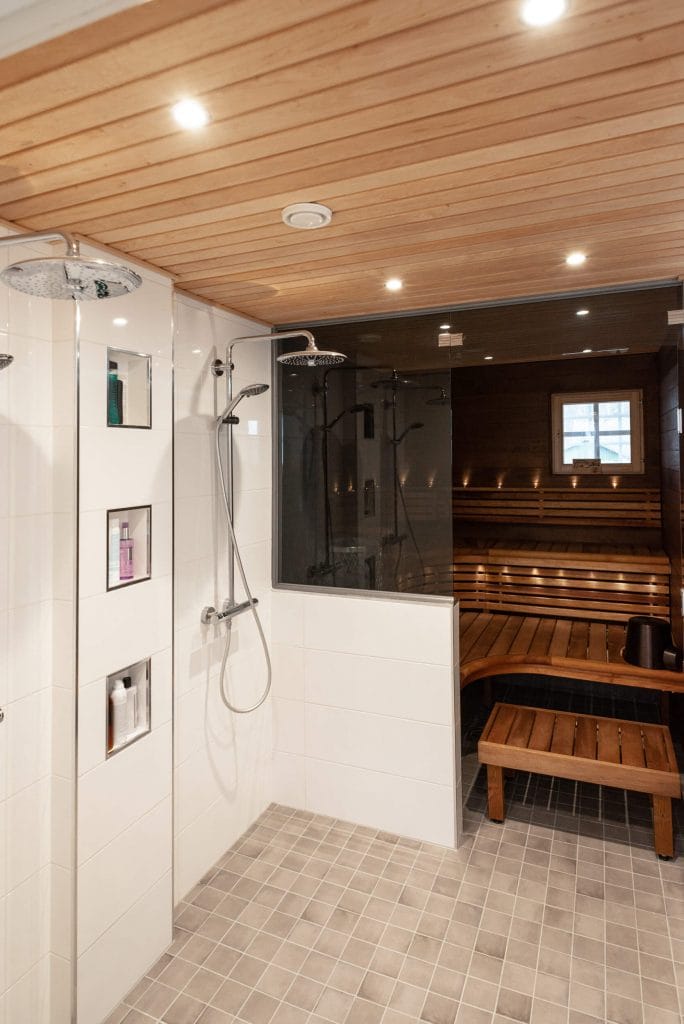 _DSC77265 - LedStore Moderni puusauna, jossa LED-valaistu kaakeloitu suihkutila, kaksi suihkupäätä, upotetut hyllyt ja lasiseinäiset lauteet pienen ikkunan vieressä.