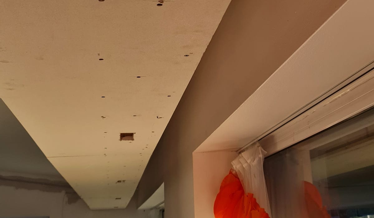 - LedStore Rakenteilla oleva katto, jossa on kipsilevyn reikiä, valotettu LED-valo ja oranssi verho ikkunan vieressä oikealla.