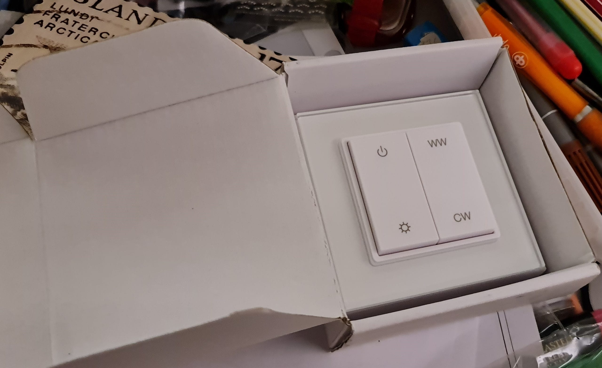 - LedStore Valkoinen valokytkin "WW"- ja "CW"-painikkeilla avoimessa laatikossa sekaisella pinnalla, jossa on värikkäitä kyniä, papereita ja pieni LED-taskulamppu.