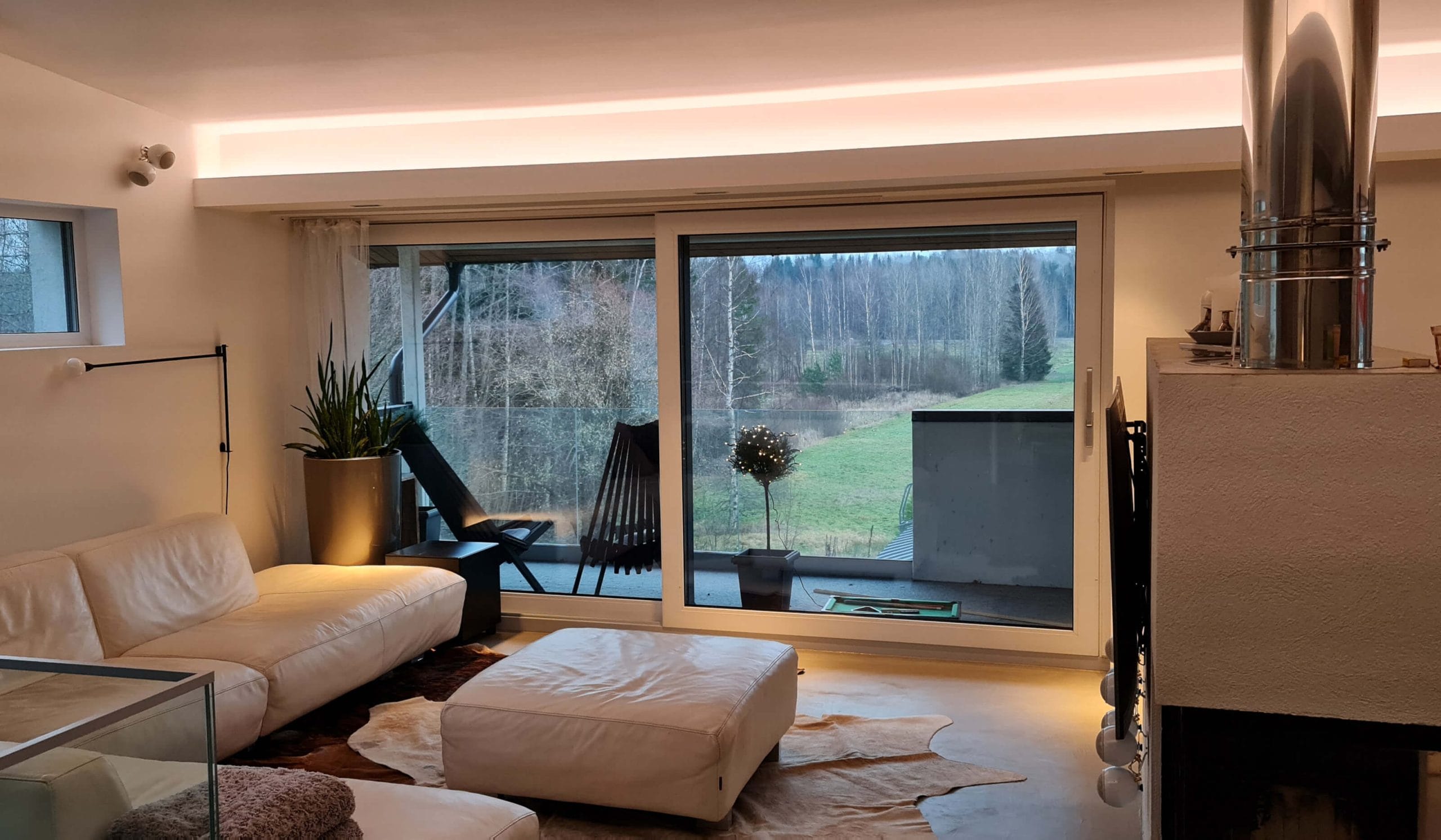 - LedStore Moderni olohuone, jossa valkoinen sohva, ottomaani, LED-valaistu lasiovet parvekkeelle; metsänäkymä; metallipiippu ja ruukkukasvit nykyaikaiseen tyyliin.