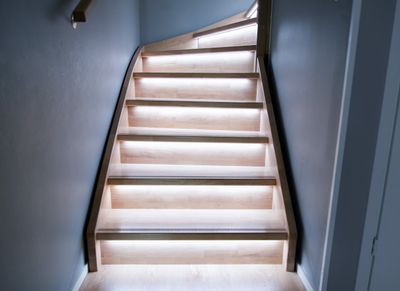 En modern trätrappa med LED-lampor under varje trappsteg, som framhäver den minimalistiska designen med varm belysning mot grå väggar.