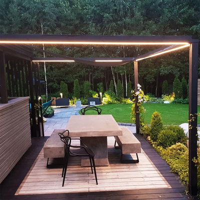 Modern uteplats med sittplatser i betong, trädäck och pergola med LED-lampor; grönskande trädgård och träd i bakgrunden.