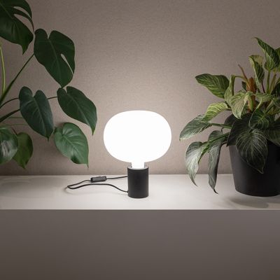 Modern LED-bordslampa med vit rund skärm på hylla, omgiven av gröna krukväxter för elegant belysning i hemmet.
