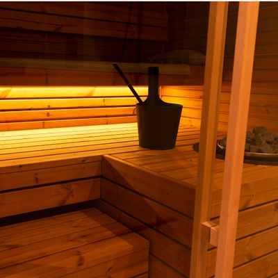Bastuinredning i trä med bänkar, mjuk LED-belysning, svart hink och skopa på bänken för en mysig och avkopplande atmosfär.