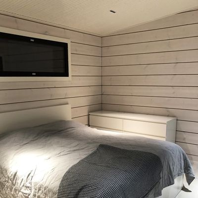 Ett mysigt sovrum med träpanel på väggarna, stämningsfull LED-belysning, grått sänglinne på sängen och en vit byrå under ett litet fönster för att skapa en lugn atmosfär.