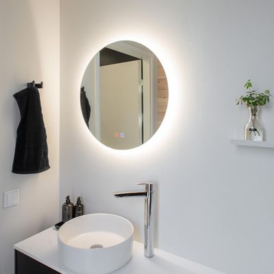 Minimalistiskt badrum med LED-belyst rund spegel, vitt handfat med kromad kran, svart handduk och liten växt på hylla.