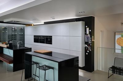 Modernt kök med högblanka vita skåp, svarta bänkskivor, köksö med barstolar, dubbelugn i rostfritt stål, kylskåp med magneter till höger under LED-belysning.