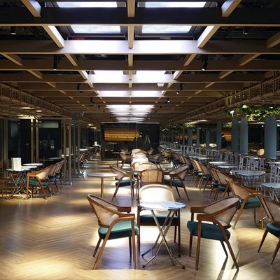 Modern inomhusrestaurang med trägolv, bord och stolar uppradade under ett gittertak med takfönster som förstärks av stämningsfull LED-belysning.