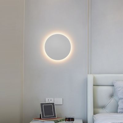 En modern cirkulär vägglampa lyser upp ett minimalistiskt sovrum med ljusgrå väggar, en vit tuftad sänggavel och en snygg sängbordsdekoration. Butik för LED-belysning.