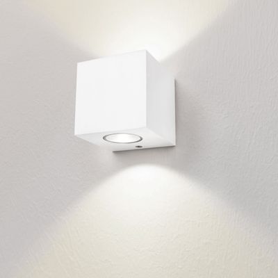 En minimalistisk vägglampa i form av en vit kub belyser en vit vägg med mjuk textur med ljus uppåt och nedåt. Perfekt för modern LED-belysning.