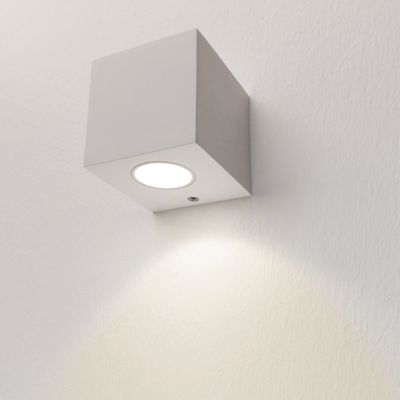 Alternativ text: kubformad LED-vägglampa som sprider mjukt vitt ljus på en strukturerad vägg, en modern belysningsdesign för ditt hem eller kontor.