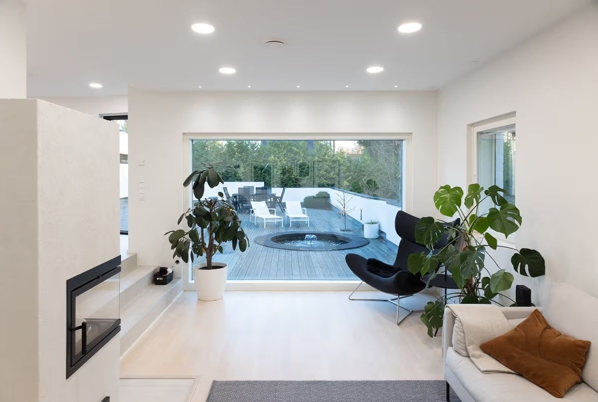 Ett vardagsrum med ljus yta och ett stort fönster med utsikt över utsidan. Det finns runda LED-paneler i taket och små spotlights framför fönstret.