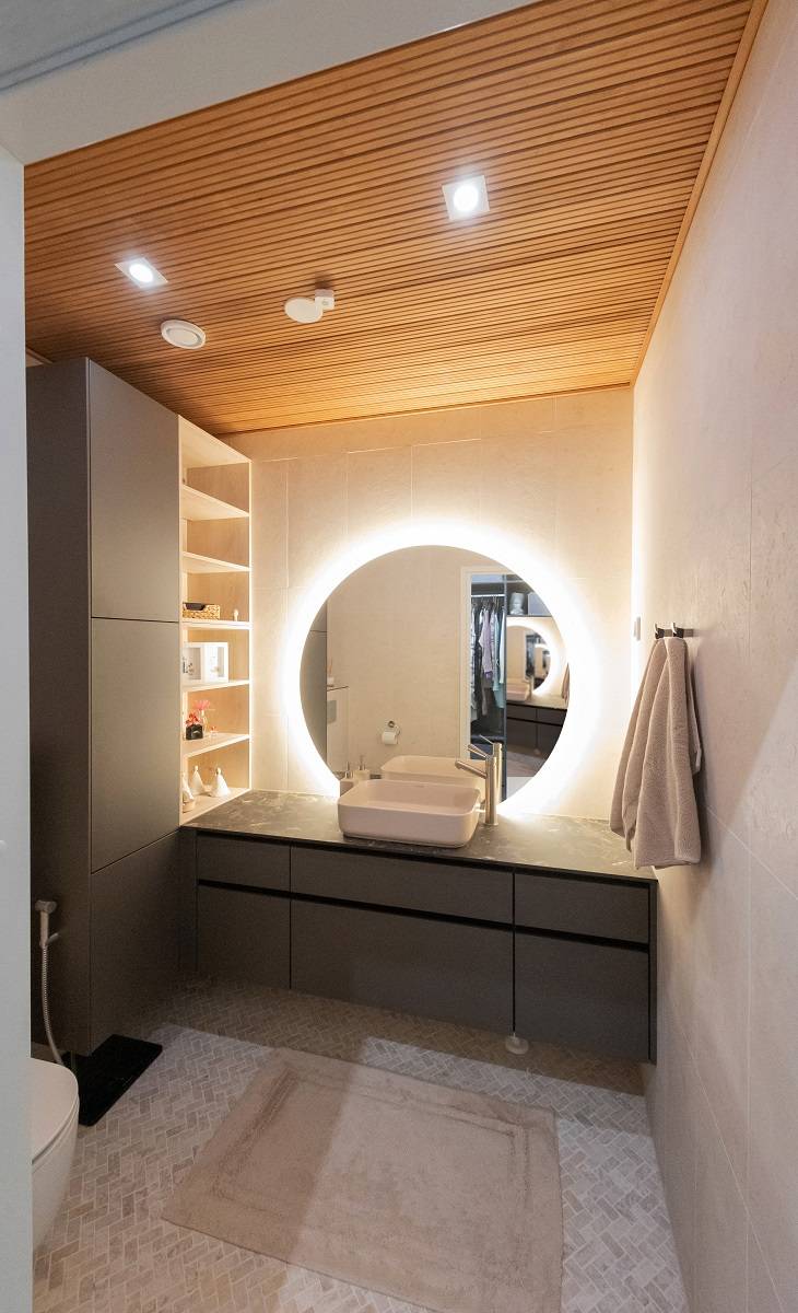 Ett modernt badrum med LED-belysning från LedStore.fi: en cirkulär LED-bakgrundsbelyst spegel ovanför handfatet skapar en elegant atmosfär. Badrummet har mörka skåp, varmt trätak och ljusa väggar. Handdukarna är placerade på höger sida och hyllorna väntar på dig på vänster sida. På golvet vilar en beige matta som bidrar till helhetsintrycket.