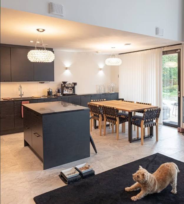 Ett modernt kök och matplats med svarta skåp, ett matbord i trä och bekväm LED-belysning från LedStore.fi skapar en mysig atmosfär. En katt som sträcker ut sig på en svart matta observerar den mysiga helheten.