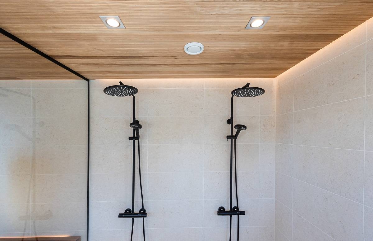 Det moderna duschrummet har två svarta duschmunstycken mot gräddfärgat kakel. Taket med träpaneler och infällda LED-lampor från LedStore.fi ger ett minimalistiskt och elegant intryck. Duschutrymmet är avskilt från resten av rummet med en yttervägg i glas.