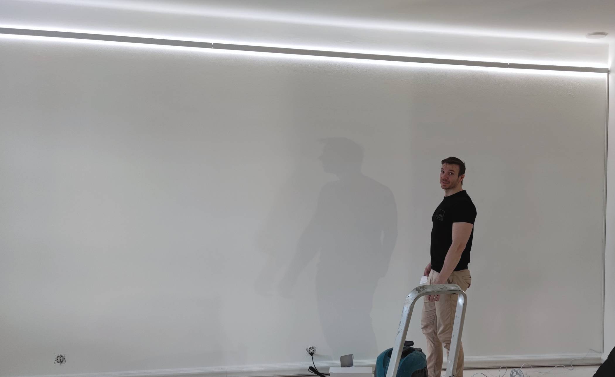 En person på en stege med ett verktyg, i mjuk LED-belysning uppifrån, mot en vit vägg. Kablar och utrustning utspridda på golvet i LED-butiksarmaturer.