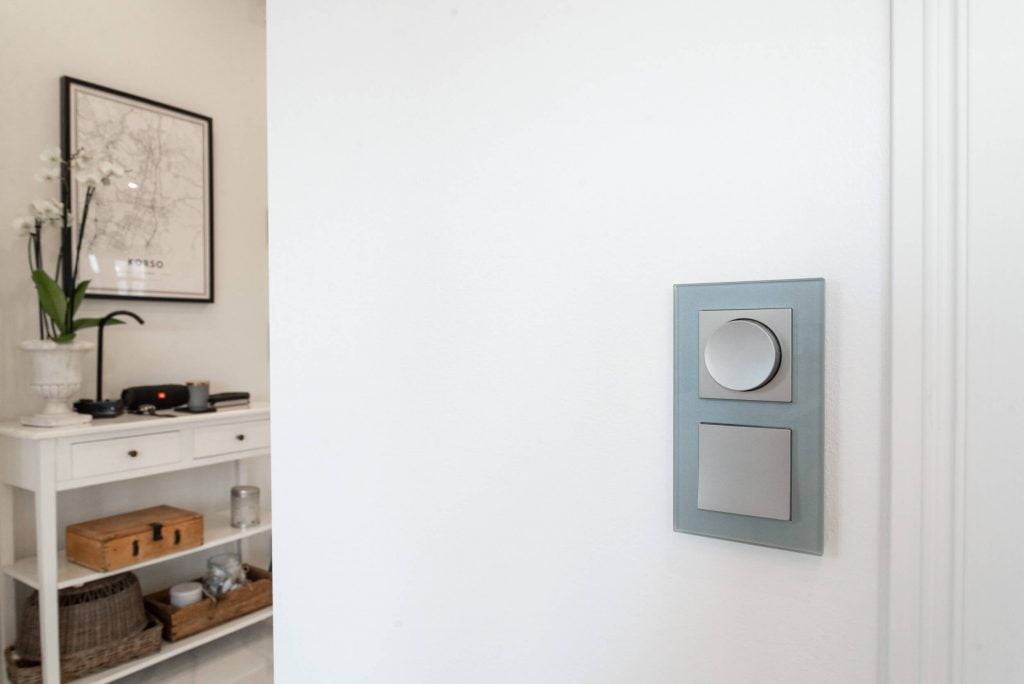 Modern LED-ljusströmbrytare med två knappar på en vit vägg; vit konsol med krukväxt och inramad karta i ett minimalistiskt rum.