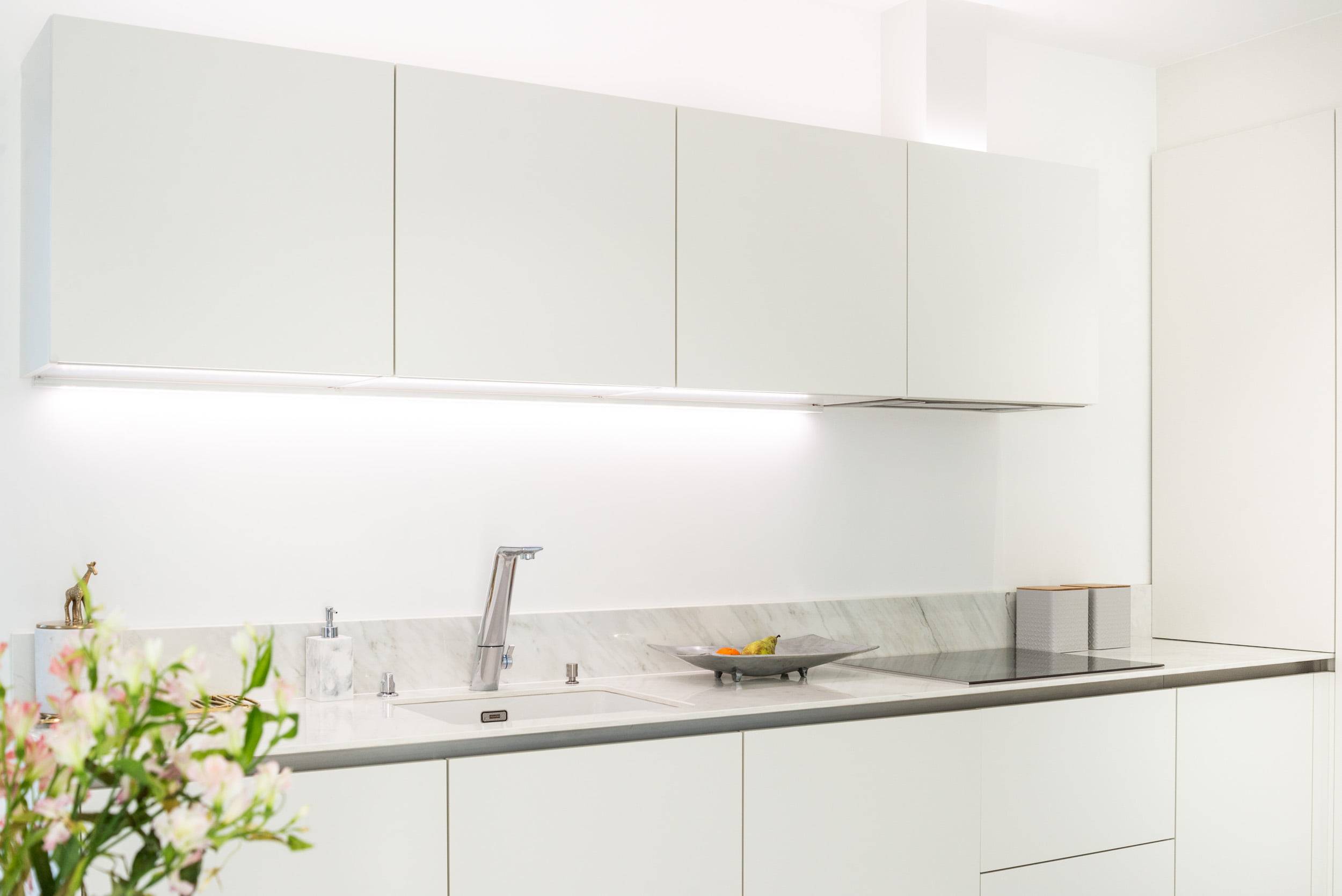 Ett stilrent kök med vita skåp, modern bänkskiva i marmor och kran i rostfritt stål. LED-belysning under bänkskivan framhäver arbetsytans utseende. En dekorativ växt och en fruktskål ger ytterligare känsla till bänkskivan. Du kan hitta rätt LED-belysningslösningar på LedStore.fi, där du kan hitta LED-produkter av hög kvalitet för att fullända utseendet på ditt kök.
