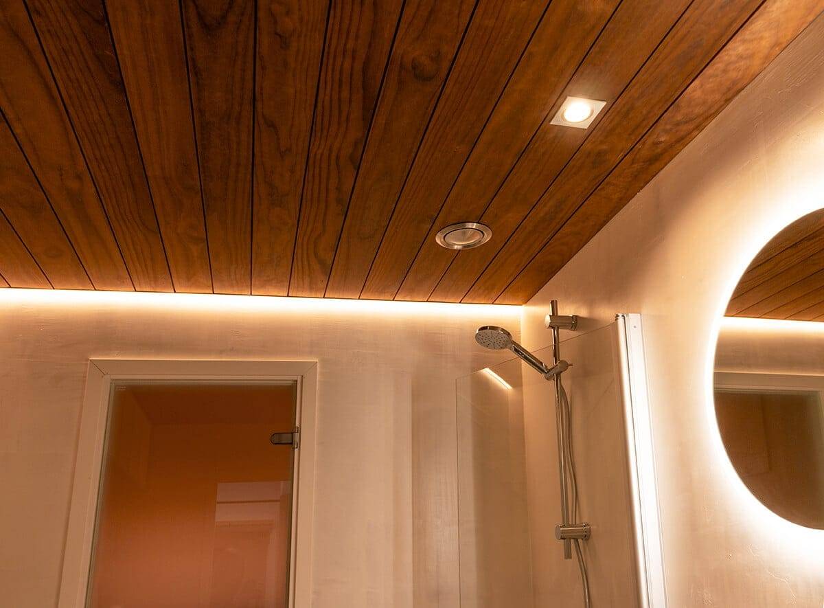 Modernissa kylpyhuoneessa på puupaneloitu katto, johon på upotettu LED-valaistus LedStore.fi:stä. Seinää koristaa pyöreä peili taustavalolla, joka sijaitsee tyylikkään suihkupään vieressä. Tämä luo tilaan lämpimän ja kutsuvan tunnelman.