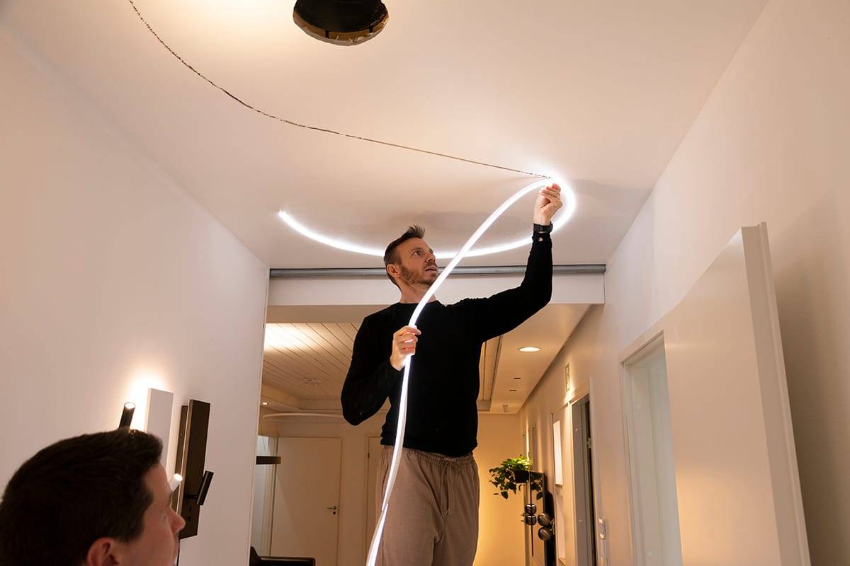 En person fäster en flexibel LED-lampremsa i ett modernt tak, vilket förbättrar belysningen i rummet. Svart tröja, beige byxor; en annan person i förgrunden, oskarp.