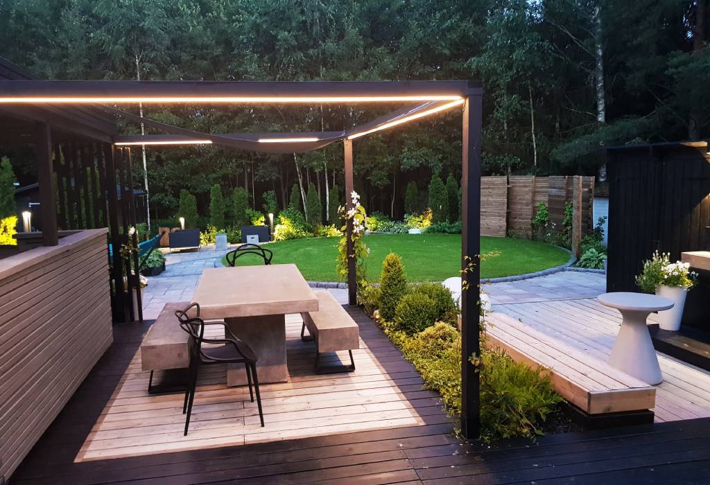 Modern utomhusterrass med pergola med inbyggd LED-belysning, betongbord, bänkar med trädäck; grönskande trädgård och välskött gräsmatta.