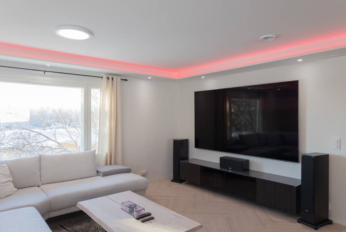 Ett modernt vardagsrum fullt av elegant atmosfär. En stor soffa inbjuder till avkoppling, medan ett soffbord i ljust trä ger rummet en varm känsla. En väggmonterad platt-TV med LED-bakgrundsbelysning ger en modern touch till rummet. Förstärk din upplevelse med golvhögtalare och njut av den röda LED-accentbelysningen i taket. Ett stort fönster släpper in naturligt ljus, vilket gör att rummet känns rymligt och ljust. Alla dessa element kombineras för att skapa den perfekta miljön med LedStore.fi:s mångsidiga LED-belysningslösningar.