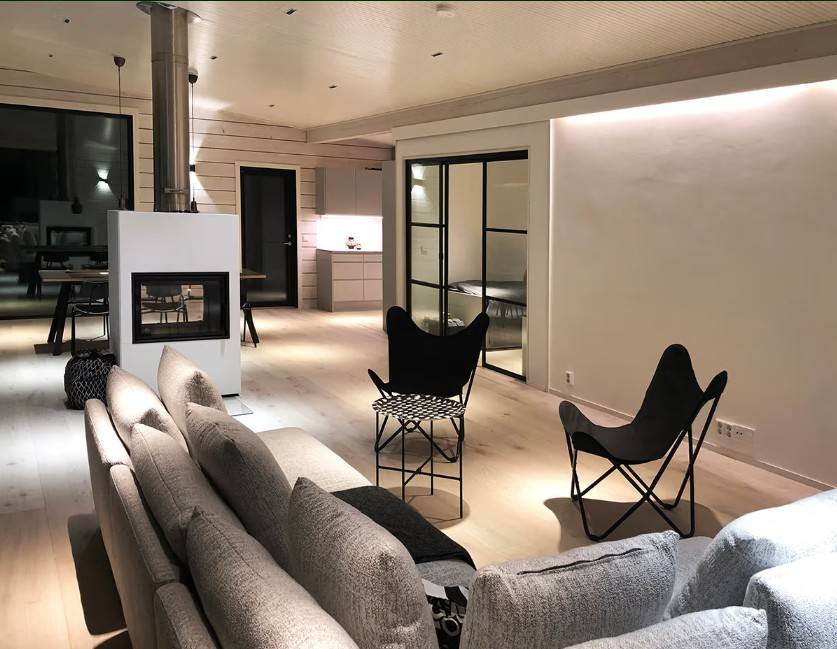 Ett modernt vardagsrum med en grå soffa, svarta stolar, en öppen spis och ett kök med öppen planlösning i bakgrunden - atmosfären och stilen skapas av LedStore.fi:s högkvalitativa LED-belysningslösning.