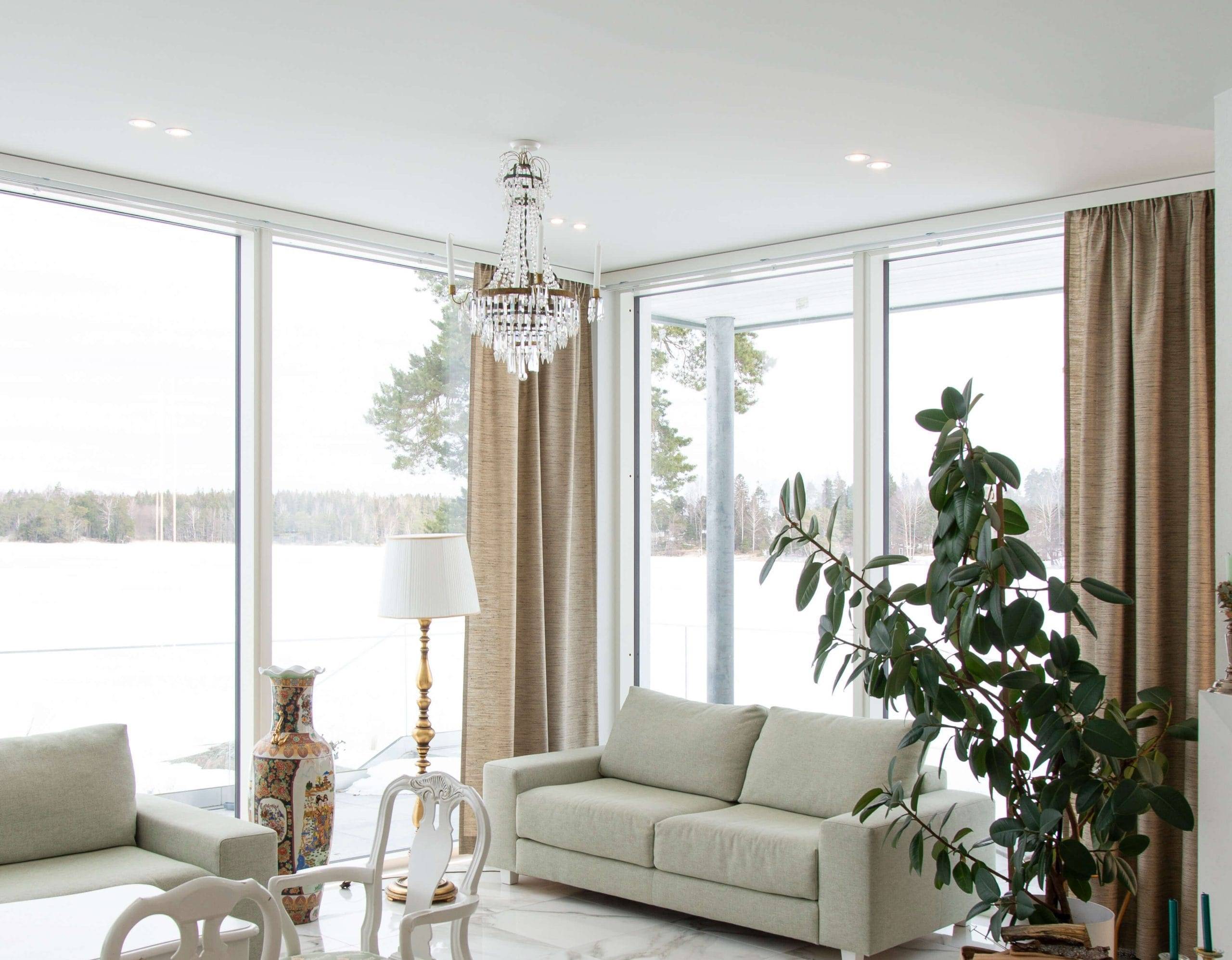 Det ljusa vardagsrummet, med stora fönster med utsikt över det snöiga landskapet, har en vit soffa, en stor grön växt och en elegant ljuskrona med LED-lampor som framhäver dess skönhet. Beige gardiner kompletterar en dekorativ vas och golvlampa, medan ett matbord med vita stolar skiner i bakgrunden. Kolla in LedStore.fi:s breda utbud av LED-armaturer och skapa en lika fantastisk belysningslösning för ditt hem!