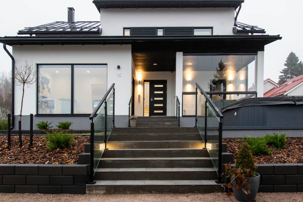 Modernt hus med vit fasad och svarta accenter; trappor med glasräcken och LED-lampor. Stora fönster, minimal landskapsarkitektur, buskar och grannhus.