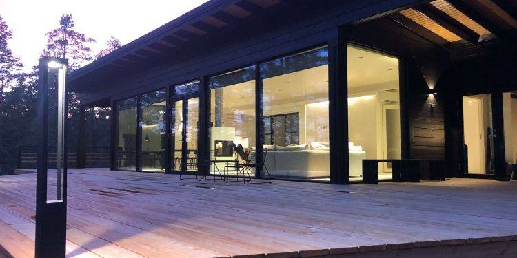 Modernt hus med stora glasfönster och rymlig träterrass, omgiven av träd. Interiören är ljust upplyst och visar minimalistisk design i handmålade toner. LED-utomhuslampor från LedStore.fi ger ett varmt sken till kvällens skymning.